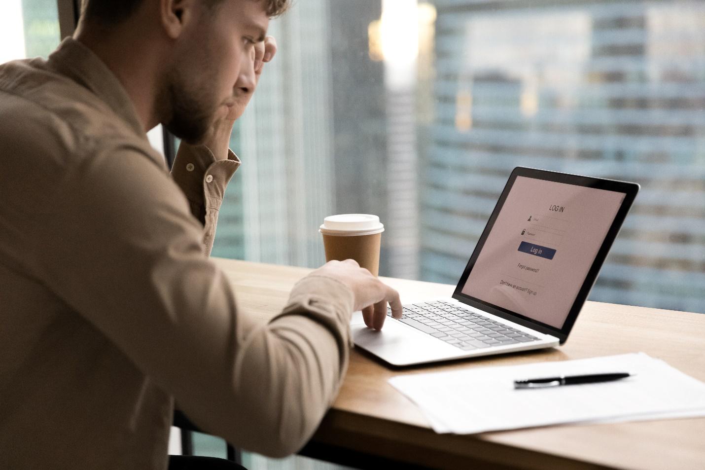 Man sitting at a desk preparing to log in to an account on his laptop with a pen and paper for notes. 