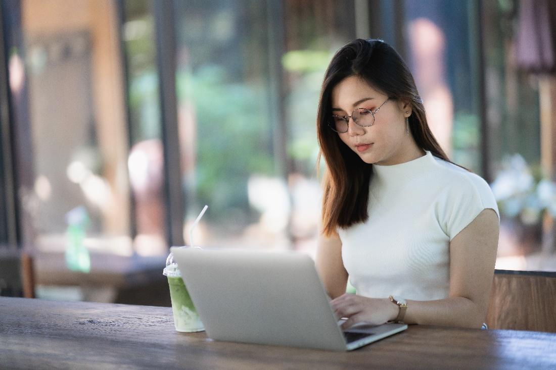 Woman wearing glasses working at a laptop with a drink nearby and a blurred background